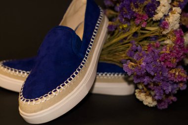 Siyah arka planda, bir çift ayakkabı, beyaz kalın tabanlı mavi süet slipler. Bir buket kuru tarla çiçeği..