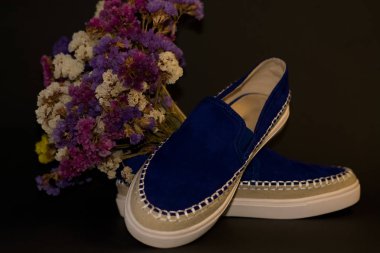 Siyah arka planda, bir çift ayakkabı, beyaz kalın tabanlı mavi süet slipler. Bir buket kuru tarla çiçeği..