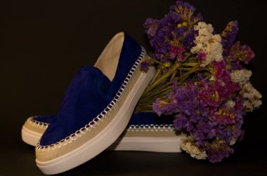 Siyah arka planda, bir çift ayakkabı, beyaz kalın tabanlı mavi süet slipler. Bir buket kuru tarla çiçeği..