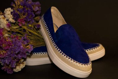 Siyah arka planda, bir çift ayakkabı, beyaz kalın tabanlı mavi süet slipler. Bir buket kuru tarla çiçeği..