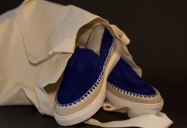 Alışveriş çantasının, alışverişçinin, bej kumaşın ve mavi süet terliklerin siyah arka planında. Sokak modası, kadın tarzı ayakkabılar..