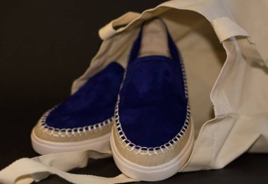 Alışveriş çantasının, alışverişçinin, bej kumaşın ve mavi süet terliklerin siyah arka planında. Sokak modası, kadın tarzı ayakkabılar..