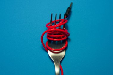 Parlak mavi arkaplanda, spagetti gibi kulaklıklardan çıkan kırmızı kabloyla çatal uzanır. Mobil aksesuarlar.