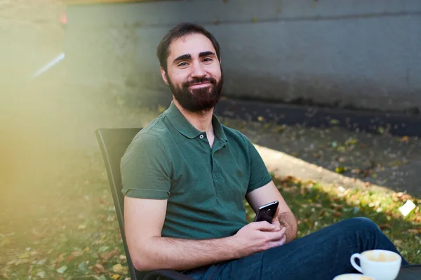 暗い髭を生やした若い男の肖像 夏のテラスでコーヒーショップに座って緑のシャツでコーヒーを飲む — ストック写真