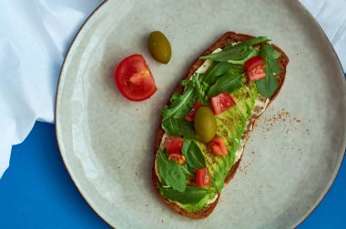 Avokado tostu, soslu çavdar ekmeği, domates, mavi arka planda büyük gri tabakta roka. Vejetaryen yararlı kahvaltı..