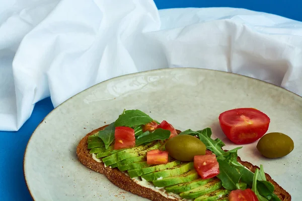 Avocado toast of rye bread with sauce, tomatoes, arugula on large grey plate on blue background. Vegetarian useful breakfast.