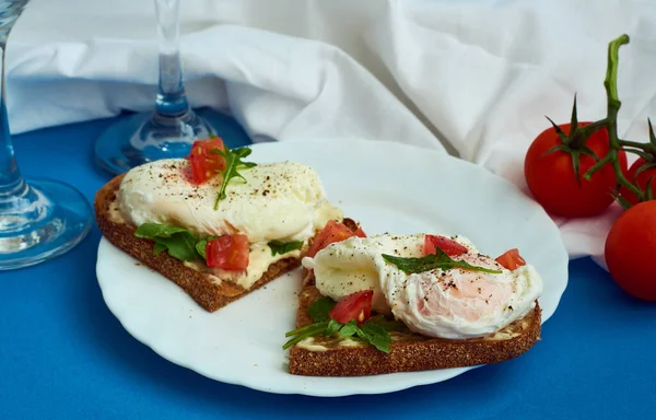 Poached eggs on sandwich of rye bread with sauce, tomatoes and arugula in white plate on blue background.