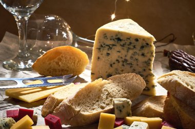 Farklı türde peynir tabağı ve parşömen parçaları, özel bıçak ve dilimlenmiş taze baget, kuru sosis çubuğu, koyu ahşap arka planda şarap bardakları..