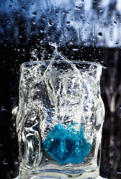 The water in the glass splashes in different directions. A glass with a crooked distorted glass with a blue ice cube falling into it, splashes of water in different directions, drops in the air.