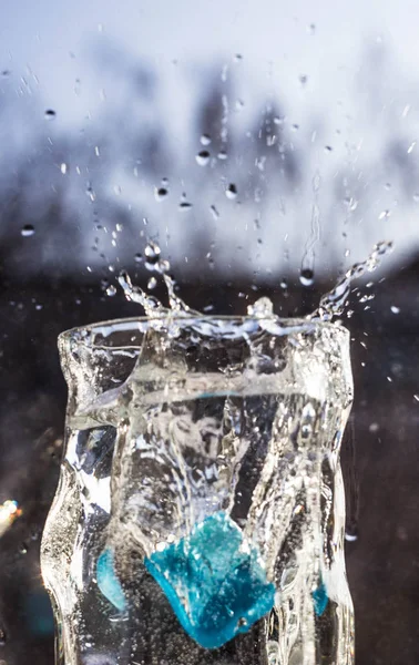 The water in the glass splashes in different directions. A glass with a crooked distorted glass with a blue ice cube falling into it, splashes of water in different directions, drops in the air.