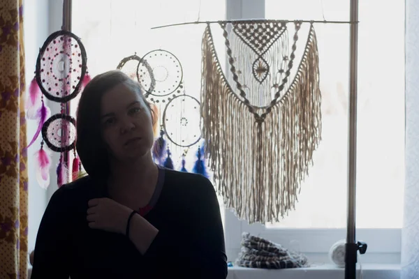 Silhouette of a woman against the window. Behind it hang dreamcatchers, macrame panels, glasses and flowers on the windowsill