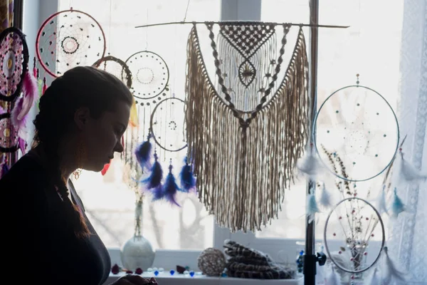 Silhouette of a woman against the window. Behind it hang dreamcatchers, macrame panels, glasses and flowers on the windowsill