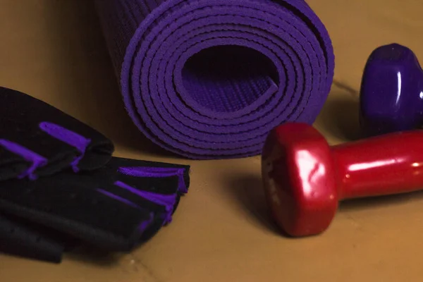 Sports equipment on the floor. On the wooden floor are fingerless gloves, two dumbbells, and a yoga Mat