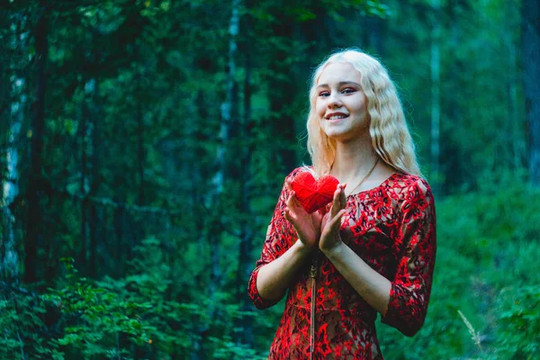 blonde in a red dress in the woods with knitted heart in hands