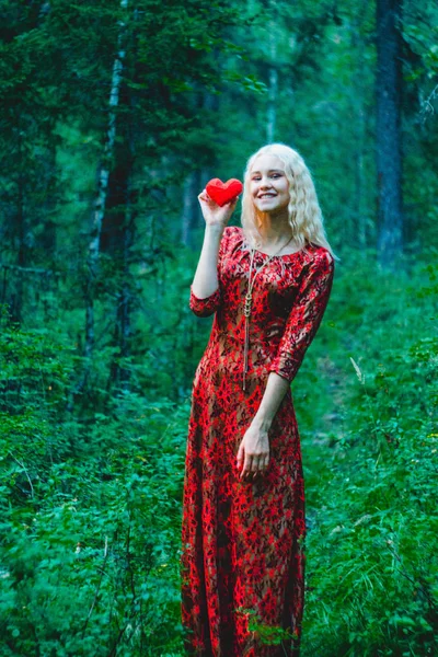 blonde in a red dress in the woods with knitted heart in hands