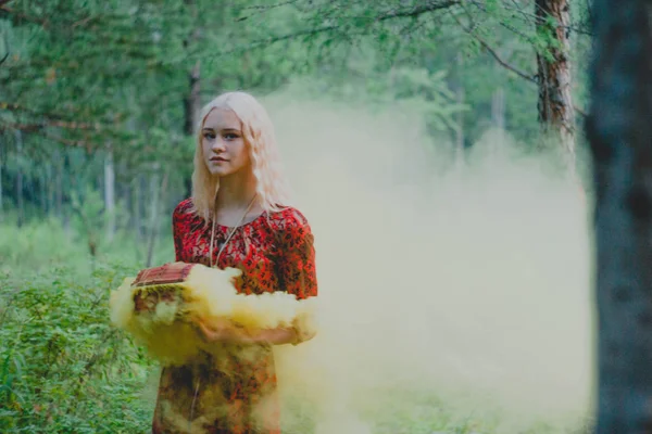 blonde in a red dress in the woods with a chest in his hands from which falls colored smoke
