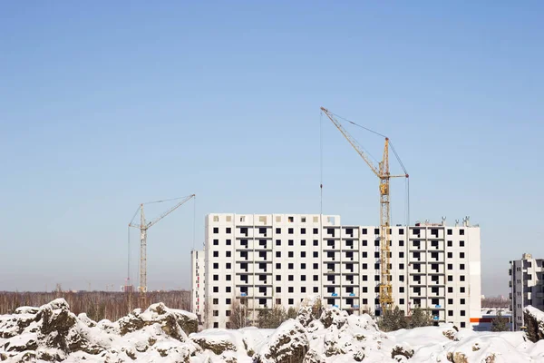 A yellow construction crane is building a house out of concrete blocks against a blue sky