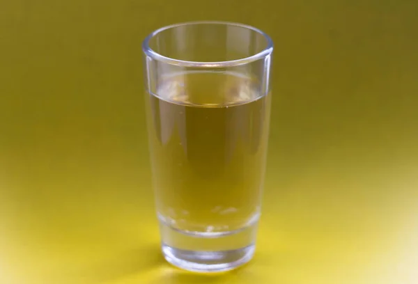 A yellow drink in a transparent glass stands on a gold background. Carbonated liquid in a shot glass. Drink in a glass