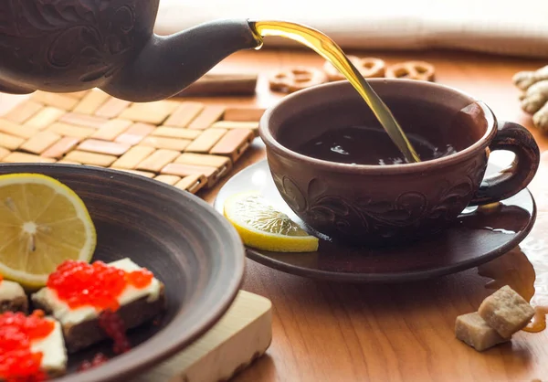 Tea is poured into a mug from the teapot. Sandwiches with butter and red caviar are on the plate.