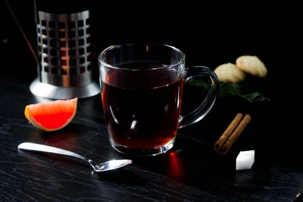 there is a clear Cup of tea on the table. a teapot, a piece of grapefruit, and a cookie in the background. The hot steam coming from the mug.