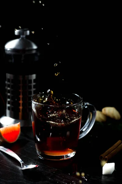 there is a clear Cup of tea on the table. a teapot, a piece of grapefruit, and a cookie in the background. The hot steam coming from the mug.