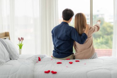 Genç ve sevimli Asyalı çiftin arka açısı yatakta kırmızı kalple birlikte oturuyorlar. Sevgililer için özel bir an için güzel zaman geçirme fikri. Evlilik teması için güzel bir konsept.