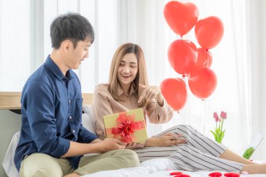 Genç ve gülümseyen Asyalı bir adam hediye paketi veriyor ve açıyor. Yatak odasında bilgisayar defteri kullanırken mutlu ve heyecanlı hisseden güzel bir kadın için hediye. Aşk fikri..