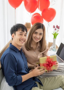 Genç ve gülümseyen Asyalı bir adam hediye paketi veriyor ve açıyor. Yatak odasında bilgisayar defteri kullanırken mutlu ve heyecanlı hisseden güzel bir kadın için hediye. Aşk fikri..