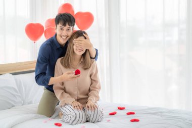 Genç ve gülümseyen Asyalı bir adam, genç ve güzel bir kadının mutlu ve heyecanlı bir yüz ifadesi takınıp, sevgilisine yüzük kutusunu açarken gözlerini kapar. Aşk kavramı ve düğün.