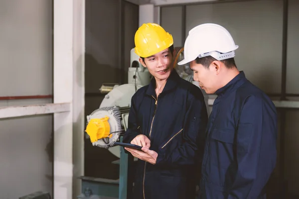 Young and handsome Asian engineers wearing dark blue coveralls and safety hard hat working together in machine room. The junior holding tablet computer and explain job details for senior boss.
