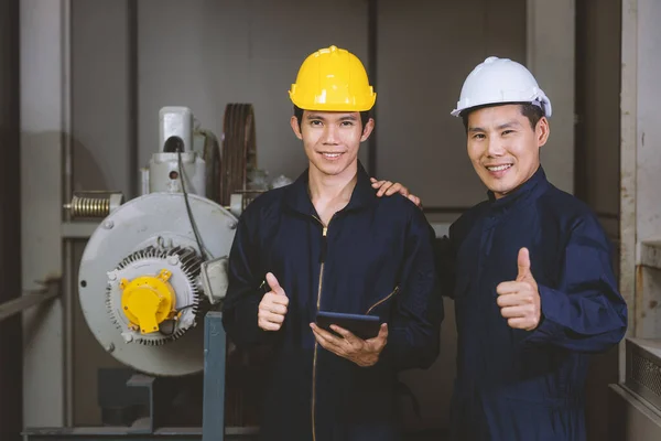 Young and handsome Asian engineers wearing dark blue coveralls and safety hard hat working together in machine room. The junior holding tablet computer and rise hands thumbs up.