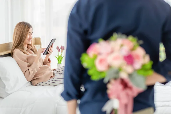 後ろにカラフルな花の男隠れて男はベッドに座ってスマートフォンを再生する女性のために与えるために準備します 女の子に焦点を当てて 愛と記念日の概念 — ストック写真