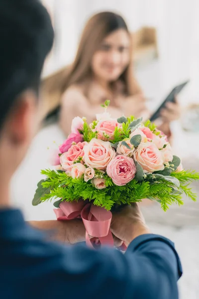 男はベッドに座ってスマートフォンを再生する女性のために与えるために準備カラフルな花の束を保持します 女の子に焦点を当てて 愛と記念日の概念 — ストック写真