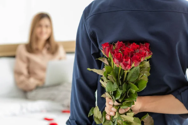 後ろにカラフルな花の束を隠して男はベッドに座ってスマートフォンを再生する女性のために与えるために準備します 女の子に焦点を当てて 愛と記念日の概念 — ストック写真