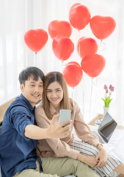 可愛いアジア系の若い女性が恋人の男の横に座り 赤い風船を背景に寝室のベッドで一緒にスマートフォンで自撮り写真を撮ります 良い瞬間のカップルのための概念 — ストック写真