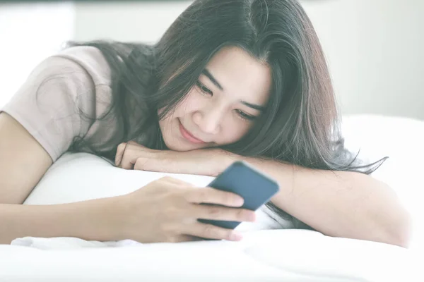 Young and beautiful woman laying on bed in bedroom in morning light and using smartphone to connect to internet with happy and relaxed manner. Idea for living alone and private time of modern woman.