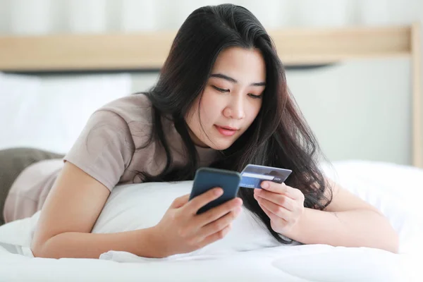 Beautiful Asian woman lying on bed in bedroom and using smartphone making shopping online payment. Mock up fake credit card to demonstrate life style of modern woman private time at home.