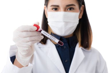 Laboratuvar önlüğü giymiş Asyalı bir kadının, elinde Covid-19 Coronavirus test tüpüyle cerrahi hijyen koruyucu maske takarken çekilmiş bulanık fotoğrafı..