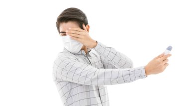 Günlük iş kıyafetleri içinde beyaz arka planda izole edilmiş tıbbi grip koruyucu maske takan genç ve yakışıklı bir adam dijital termometrenin okumasından sonra heyecanlanıyor, geriliyor ve korkuyor..