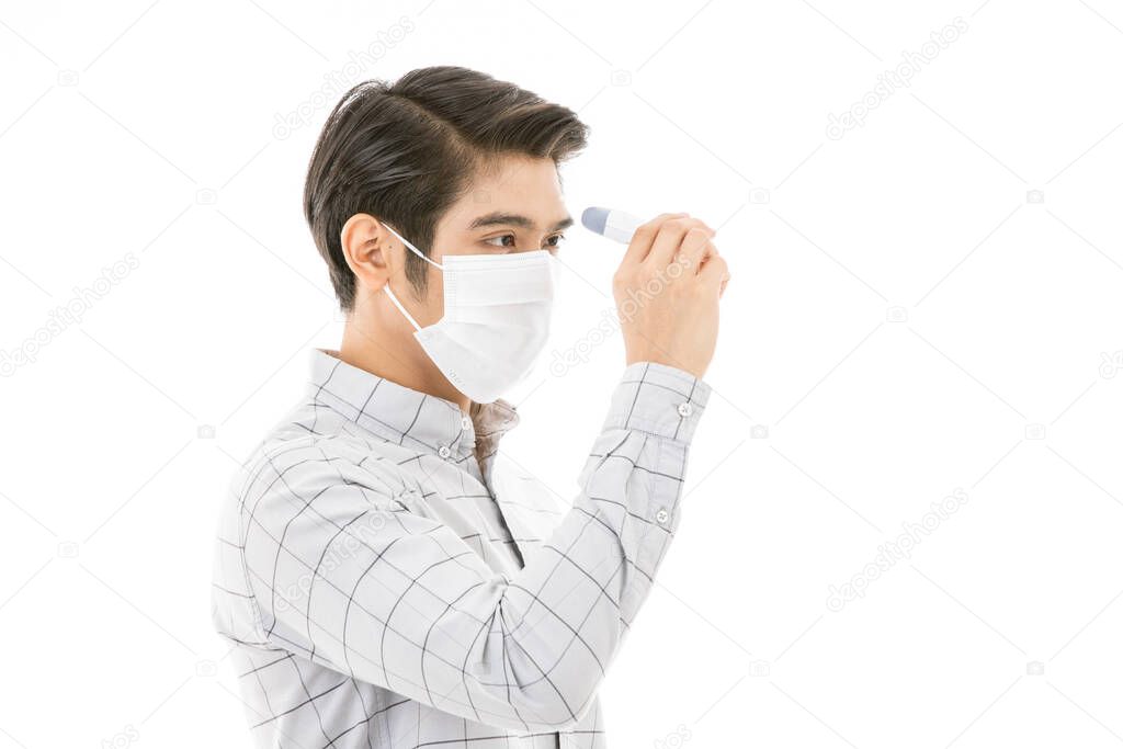 Young and handsome man in casual business cloth wearing medical flu protective mask isolated on white background. using digital infrared thermometer to measure and check himself at forehead.