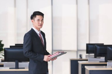 Yakışıklı Asyalı işadamı gülümsüyor, ayakta duruyor ve elinde bilgisayarla ofisinde duruyor..