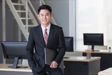 Yakışıklı Asyalı işadamı gülümsüyor, ayakta duruyor ve elinde bilgisayarla ofisinde duruyor..