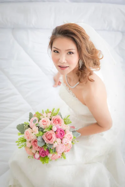 Portrait of beautiful Asian bride sitting in the dressing room and holding a bouquet of flowers .