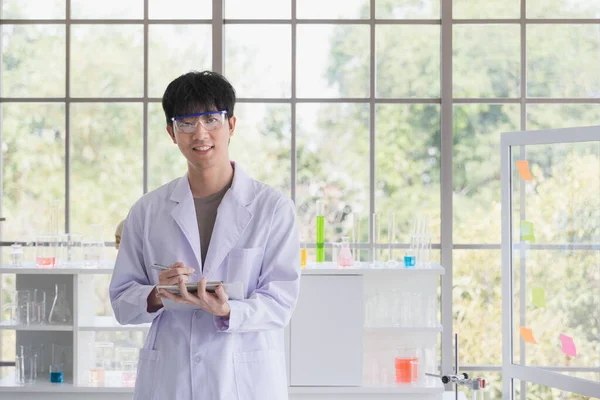 Asian male scientist standing, poses and look at camera in laboratory room, colorful beakers on shelf behind him.