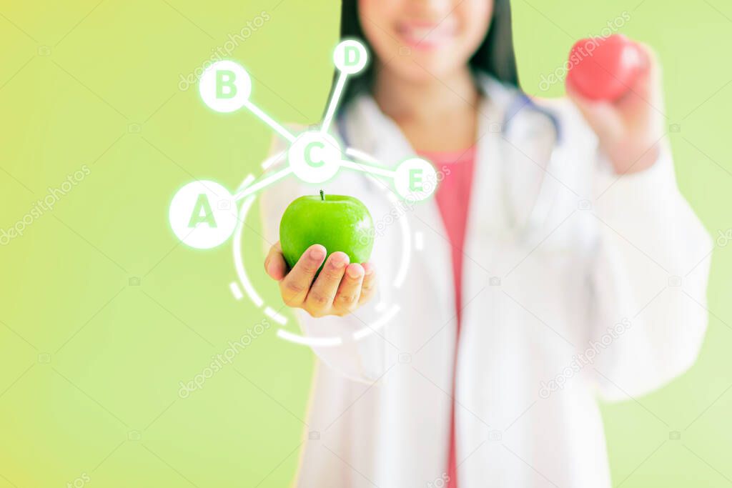 Close up of double exposure of blurred beautiful Asian female doctor hold apple with fruit-based vitamin icons such as A, B, C, D, E, all of it that derived from nature isolated on green background.