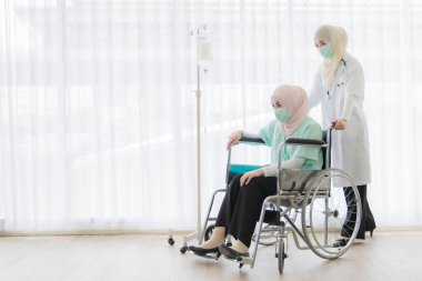 Tesettürlü Müslüman kadın doktor hastanede tekerlekli sandalyede oturan hastaya nazikçe yardım ediyor. Virüs ve hastalıklardan korunmak için cerrahi hijyen maskesi takıyorlar..