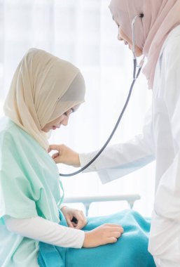 Yüzüne hijyen koruması maskesi takan Müslüman bir kadın doktor hastane odasında, stetoskop kullanarak, kadın hastayla kibarca ilgileniyor ve onunla dikkatlice konuşuyor..