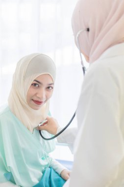 Yüzüne hijyen koruması maskesi takan Müslüman bir kadın doktor hastane odasında, stetoskop kullanarak, kadın hastayla kibarca ilgileniyor ve onunla dikkatlice konuşuyor..