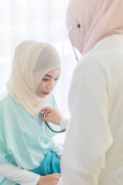 Yüzüne hijyen koruması maskesi takan Müslüman bir kadın doktor hastane odasında, stetoskop kullanarak, kadın hastayla kibarca ilgileniyor ve onunla dikkatlice konuşuyor..