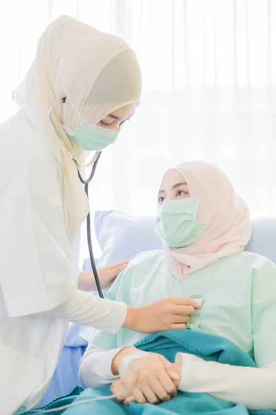 顔に衛生保護外科用マスクを身に着けているイスラム教徒の女性医師は 慎重に聴診器を使用して 病院の部屋で優しさと女性患者にケアと話をチェック — ストック写真